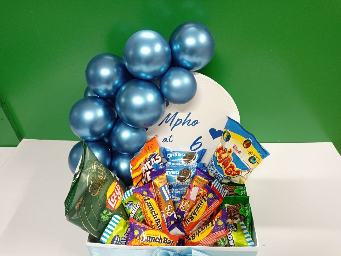 Gift basket with blue balloons, chocolate bars, and on a white surface against a green background