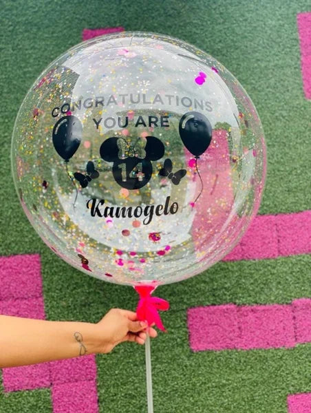 Clear balloon with Mickey Mouse design and text held by a person on a pink and green checkered background