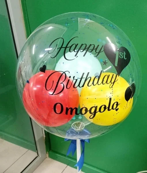Clear balloon with red and yellow balloons inside, displaying 'Happy 1st Birthday Omogolo' against a green door.