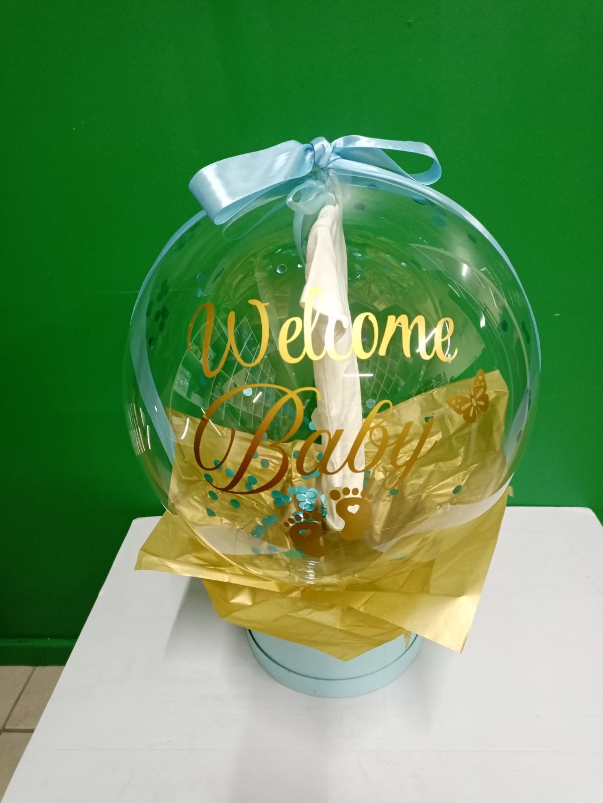 Clear decorative ball with 'Welcome Baby' text, gold accents, and a blue ribbon against a green background.  Gifting in Pretoria.