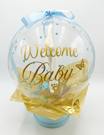 Clear balloon with 'Welcome Baby' text, blue ribbon, and gold accents on a white background