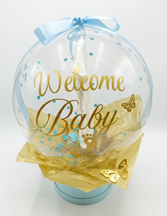 Clear balloon with 'Welcome Baby' text, blue ribbon, and gold accents on a white background
