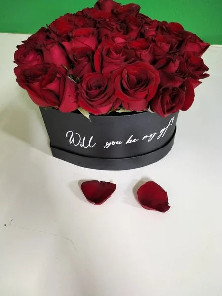 Black box with red roses and 'Will you be my gf?' text, on a white surface with green background