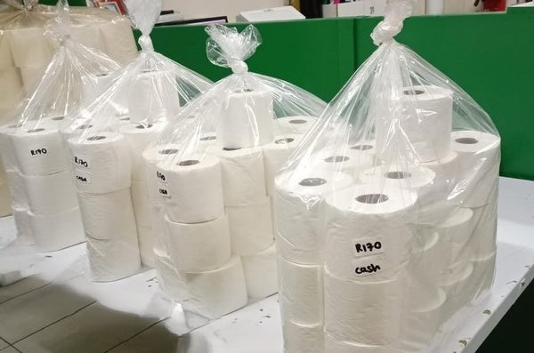 Multiple rolls of toilet paper in plastic bags on a table with a green wall in the background.