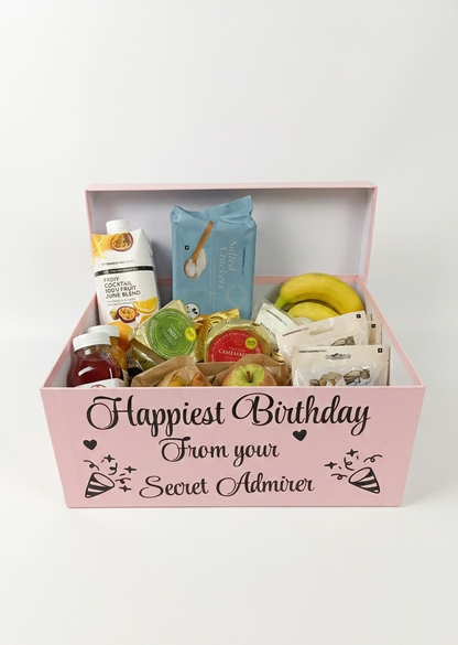 Gift basket with various items including snacks and a bottle, with 'Happiest Birthday From your Secret Admirer' text on a pink box.