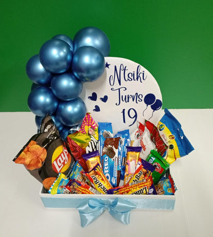 Box of assorted snacks with blue balloons and a personalized sign 
